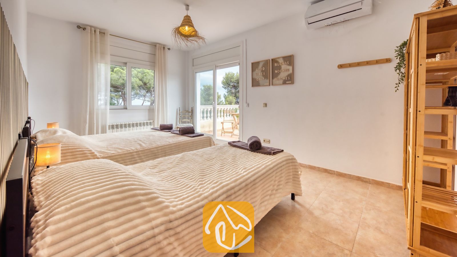 Vakantievilla's Costa Brava Spanje - Villa Ibiza - Slaapkamer met twee eenpersoonsbedden voorzien van gestructureerde, beige dekens, een houten wandrek en een glazen deur die uitkomt op een balkon met uitzicht op groene bomen.