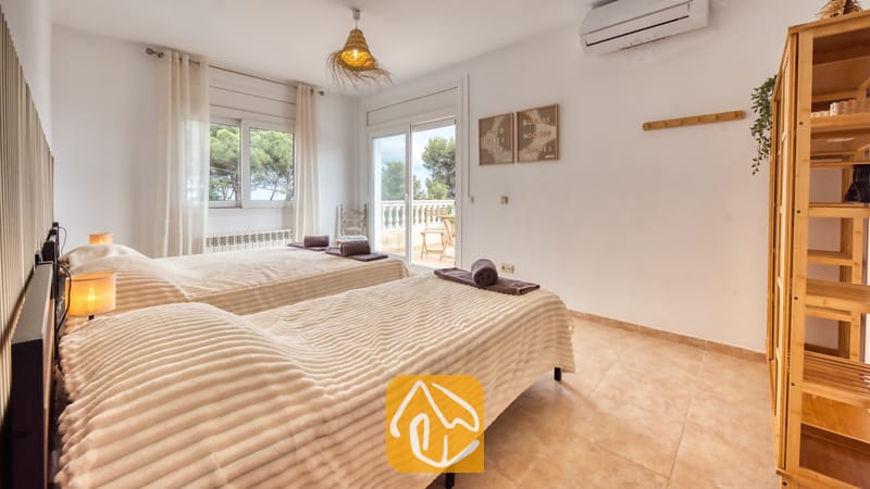 Villas de vacances Costa Brava Espagne - Villa Ibiza - Chambre avec deux lits jumeaux ornés de couvertures texturées beige, une étagère en bois et une porte vitrée s'ouvrant sur un balcon avec vue sur les arbres verdoyants à l'extérieur.