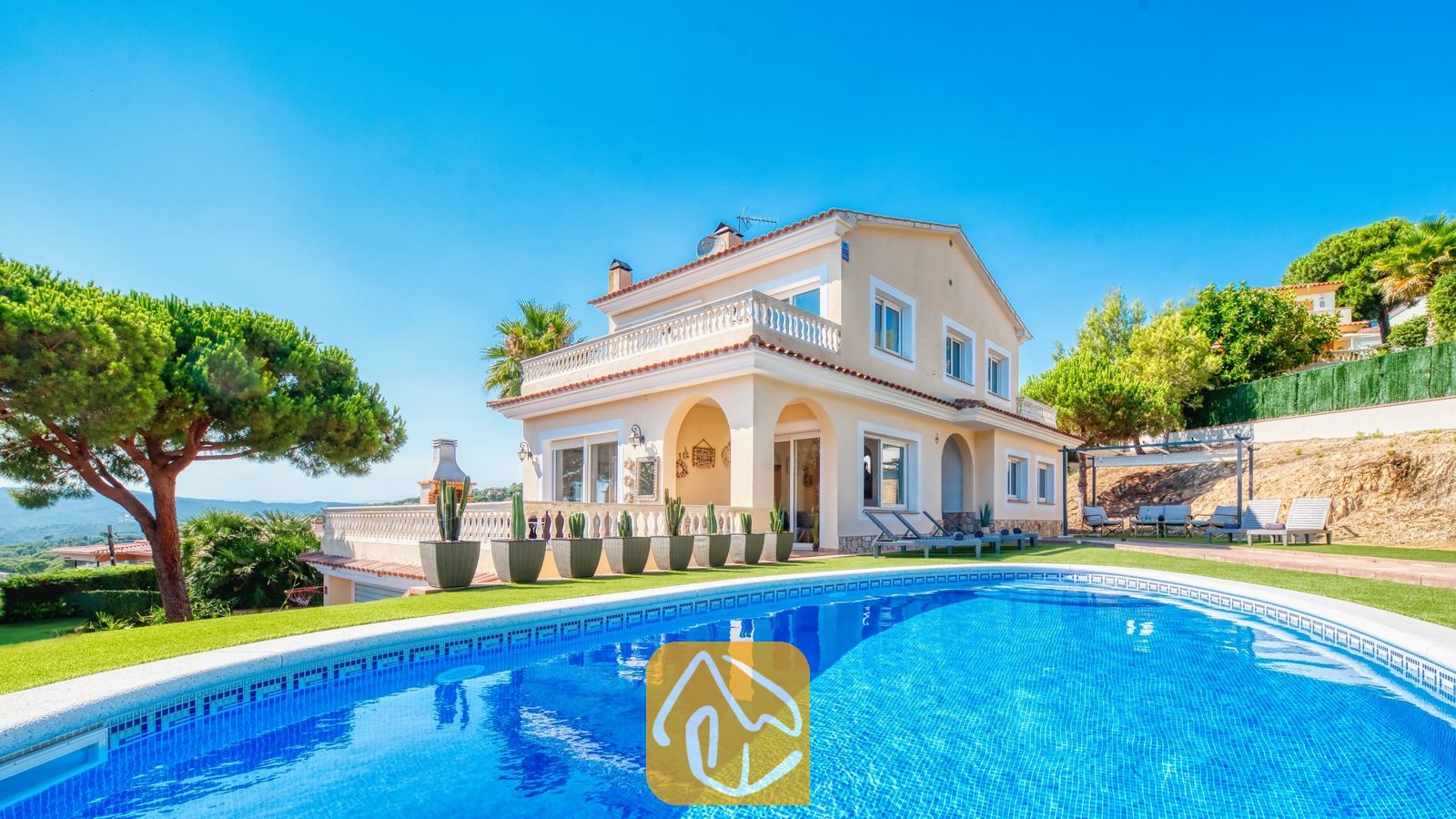 Vakantievilla's Costa Brava Spanje - Villa Ibiza - Privézwembad op de voorgrond met Villa Ibiza, een crèmekleurig huis van twee verdiepingen, een groen gazon en cactussen in potten onder een helderblauwe hemel.