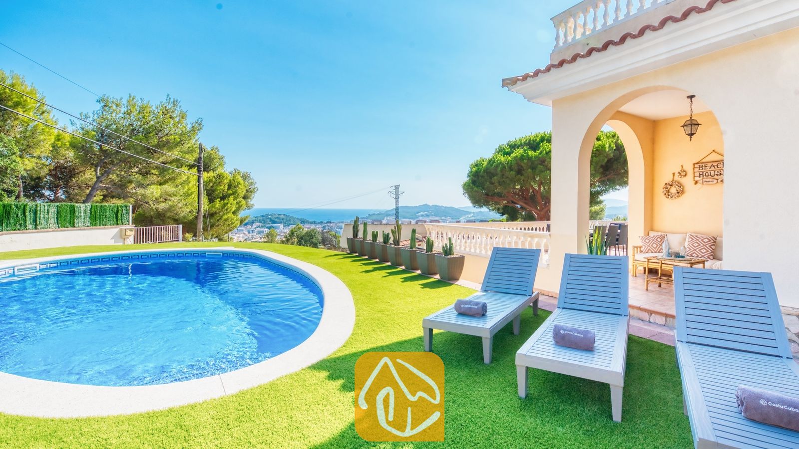 Vakantievilla's Costa Brava Spanje - Villa Ibiza - Privézwembad en blauwe houten ligstoelen op kunstgras bij Villa Ibiza met uitzicht op de verre zee en bomen.