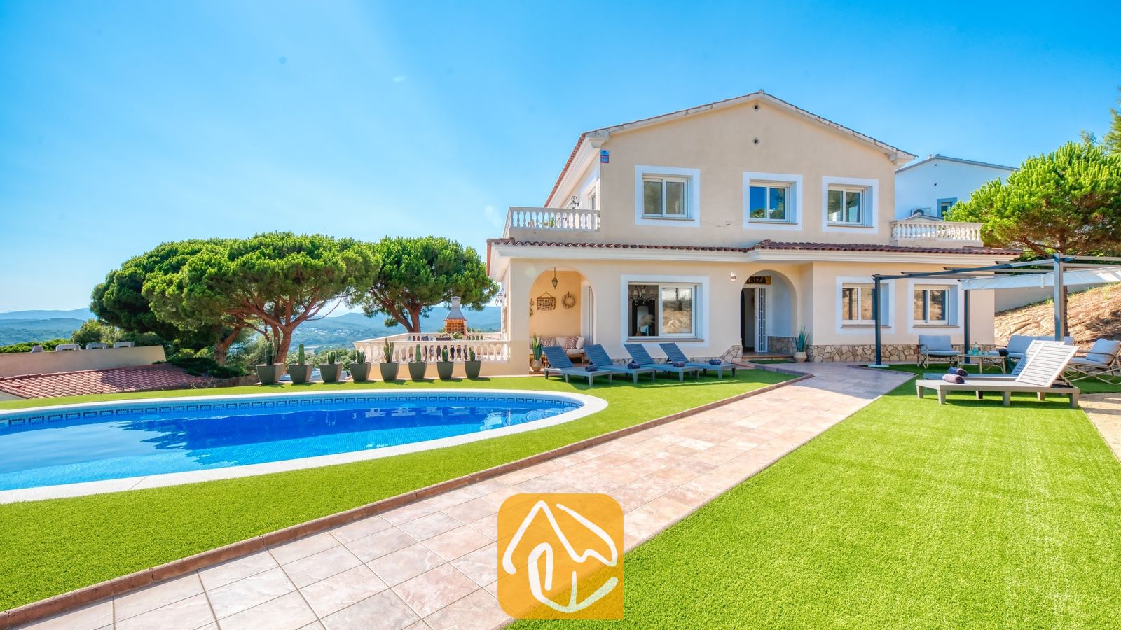 Vakantievilla's Costa Brava Spanje - Villa Ibiza - Buitenaanzicht van Villa Ibiza met een gebogen blauw zwembad, groen kunstgras, ligbedden en een beige huis van twee verdiepingen onder een helderblauwe hemel