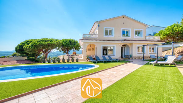 Vakantievilla's Costa Brava Spanje - Villa Ibiza - Buitenaanzicht van Villa Ibiza met een gebogen blauw zwembad, groen kunstgras, ligbedden en een beige huis van twee verdiepingen onder een helderblauwe hemel