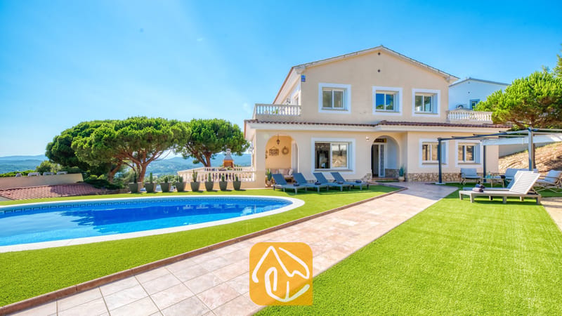 Vakantievilla's Costa Brava Spanje - Villa Ibiza - Buitenaanzicht van Villa Ibiza met een gebogen blauw zwembad, groen kunstgras, ligbedden en een beige huis van twee verdiepingen onder een helderblauwe hemel
