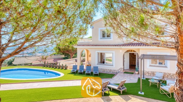 Vakantievilla's Costa Brava Spanje - Villa Ibiza - Buitenaanzicht van Villa Ibiza met een gebogen zwembad, een kunstgrasveld, meerdere grijze ligbedden en een beige huis van twee verdiepingen met terracotta dakpannen.