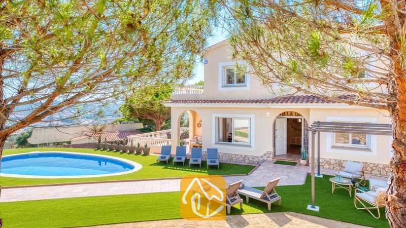 Vakantievilla's Costa Brava Spanje - Villa Ibiza - Buitenaanzicht van Villa Ibiza met een gebogen zwembad, een kunstgrasveld, meerdere grijze ligbedden en een beige huis van twee verdiepingen met terracotta dakpannen.