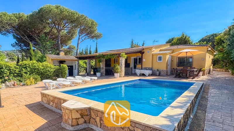 Vakantievilla's Costa Brava Spanje - Villa Iako - Het privézwembad en de gele buitenzijde van Villa Iako, voorzien van stenen bestrating, ligstoelen en een eethoek buiten onder een parasol tegen een helderblauwe hemel.