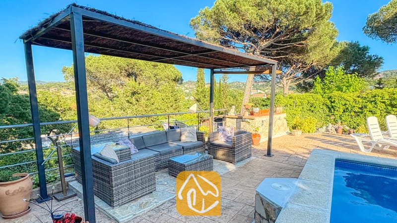 Vakantievilla's Costa Brava Spanje - Villa Iako - Buitenterras van Villa Iako met een grijze ritzel-loungeset onder een donkere metalen pergola, gelegen naast een zwembad met stenen tegels en uitzicht op de groene heuvels.