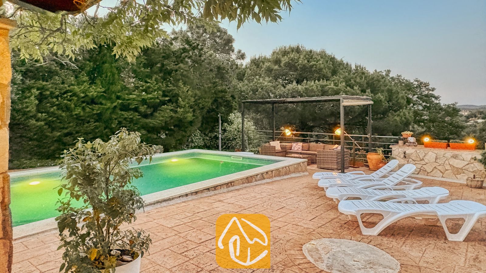 Vakantievilla's Costa Brava Spanje - Villa Iako - Buiten terras van Villa Iako met een verlicht zwembad, witte kunststof ligstoelen en een overdekt zitgedeelte omringd door bomen.