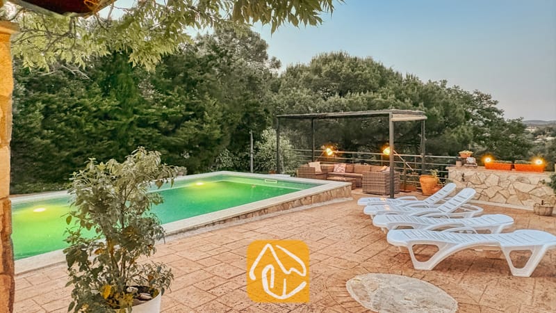 Villas de vacances Costa Brava Espagne - Villa Iako - Terrasse extérieure de la Villa Iako dotée d'une piscine éclairée, de bains de soleil blancs en plastique et d'un coin salon ombragé entouré d'arbres.