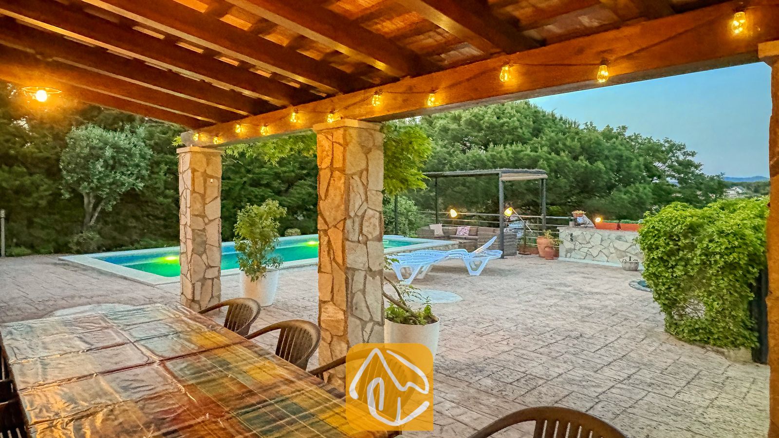 Vakantievilla's Costa Brava Spanje - Villa Iako - Overdekt buiten eetterras bij Villa Iako met een balkenplafond van hout, sfeerverlichting en stenen zuilen met uitzicht op een zwembad en bomen.