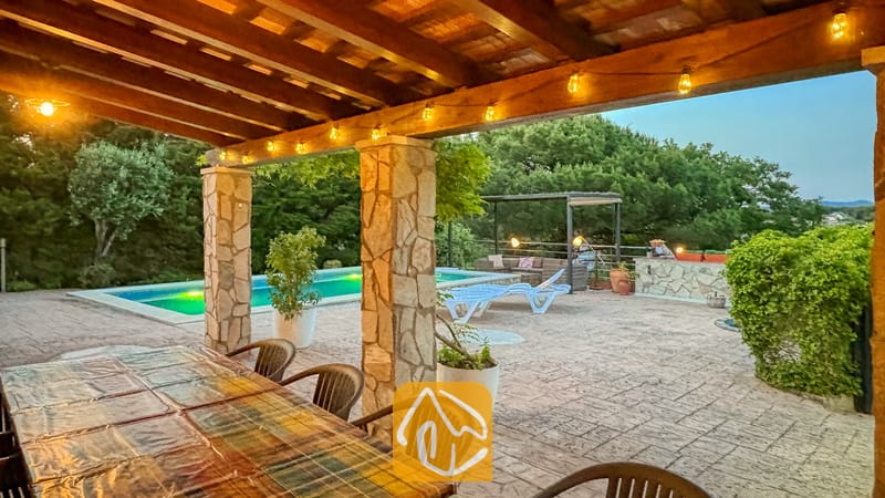 Vakantievilla's Costa Brava Spanje - Villa Iako - Overdekt buiten eetterras bij Villa Iako met een balkenplafond van hout, sfeerverlichting en stenen zuilen met uitzicht op een zwembad en bomen.