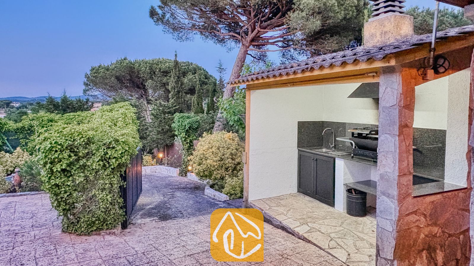 Vakantievilla's Costa Brava Spanje - Villa Iako - Buitenkeuken met barbecue en stenen schoorsteen, donkere kasten en een spoelbak, gelegen naast een verharde oprijlaan bij Villa Iako.