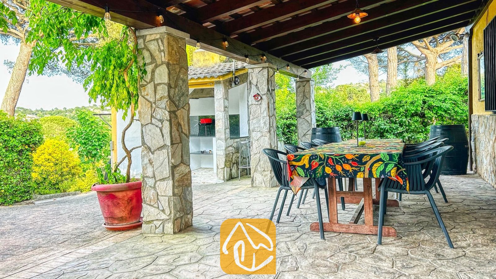 Vakantievilla's Costa Brava Spanje - Villa Iako - Overdekt buitenterras van Villa Iako met een grote houten tafel, een tafelkleed met motief, stenen pilaren en een ingebouwde barbecue op de achtergrond.