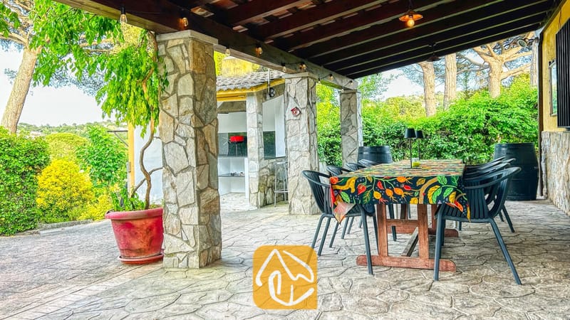 Vakantievilla's Costa Brava Spanje - Villa Iako - Overdekt buitenterras van Villa Iako met een grote houten tafel, een tafelkleed met motief, stenen pilaren en een ingebouwde barbecue op de achtergrond.