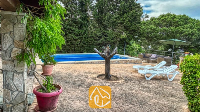 Vakantievilla's Costa Brava Spanje - Villa Iako - Villa Iako buitenterras met een rechthoekig zwembad, witte kunststof ligstoelen en terracotta bloempotten tegen een achtergrond van dichte, groene bomen.
