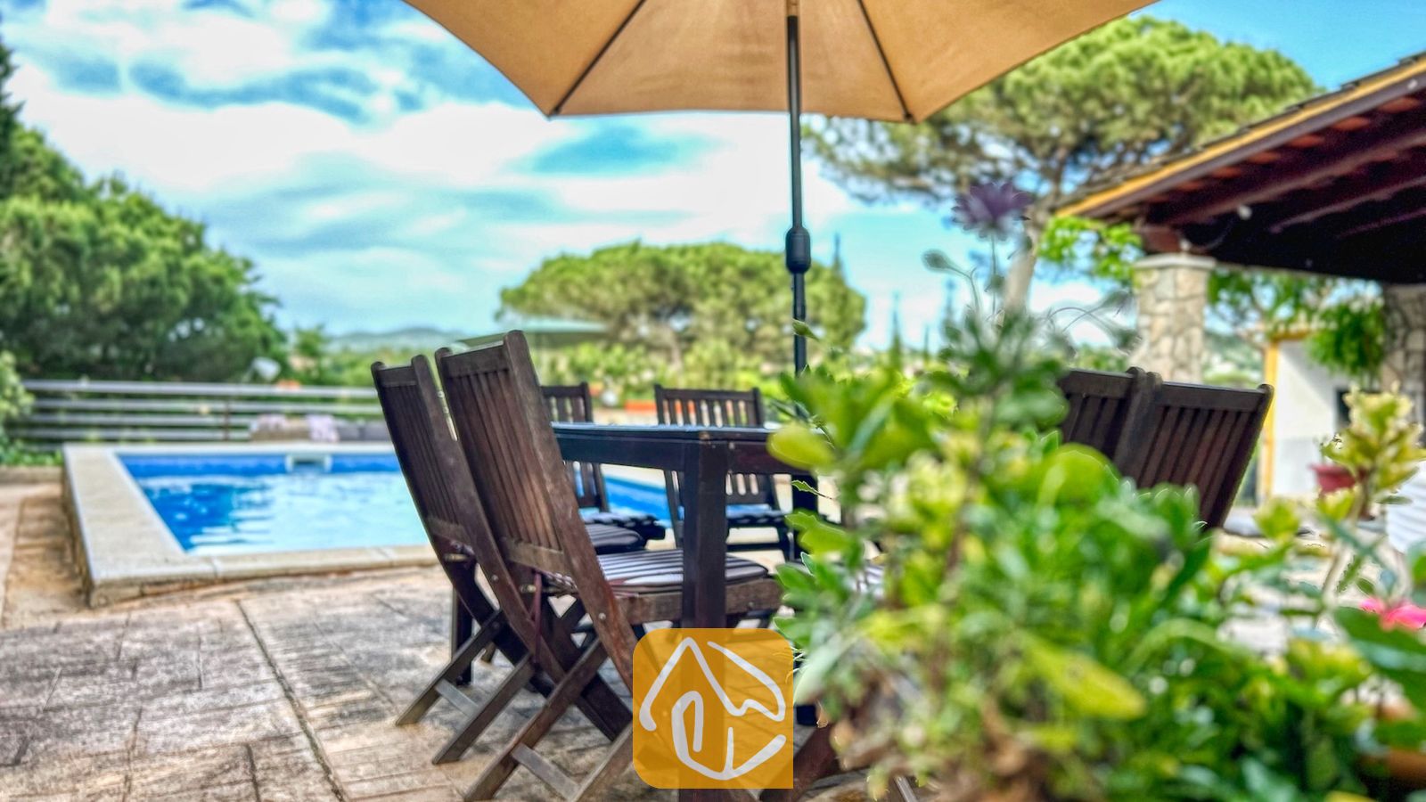 Vakantievilla's Costa Brava Spanje - Villa Iako - Houten eethoek onder een parasol naast het zwembad van Villa Iako, met groene bomen op de achtergrond.