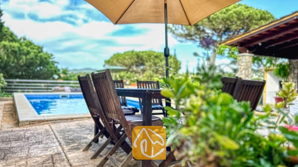 Vakantievilla's Costa Brava Spanje - Villa Iako - Houten eethoek onder een parasol naast het zwembad van Villa Iako, met groene bomen op de achtergrond.