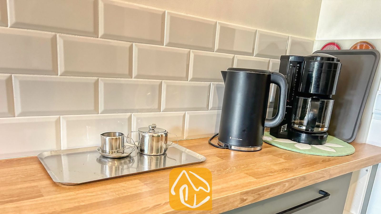 Vakantievilla's Costa Brava Spanje - Villa Iako - Zwarte elektrische waterkoker en koffiezetapparaat op een houten aanrechtblad tegen een beige tegelachterwand, met een klein zilveren dienblad met metalen kopjes.
