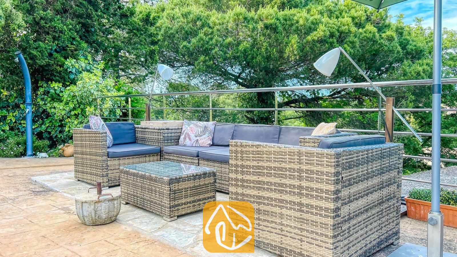 Vakantievilla's Costa Brava Spanje - Villa Iako - Buiten loungeset van rotan met donkere kussens en sierkussens op een stenen terras bij Villa Iako, omringd door bomen.