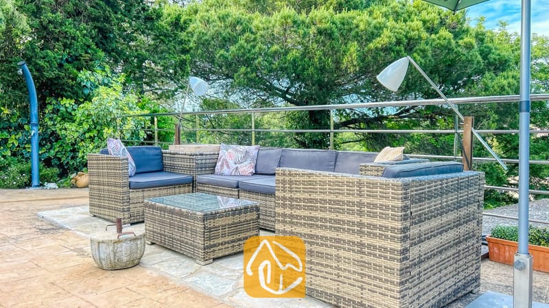 Vakantievilla's Costa Brava Spanje - Villa Iako - Buiten loungeset van rotan met donkere kussens en sierkussens op een stenen terras bij Villa Iako, omringd door bomen.