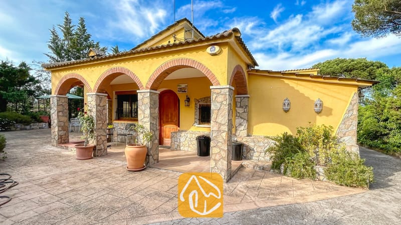 Vakantievilla's Costa Brava Spanje - Villa Iako - Gele buitenkant van Villa Iako met een overdekt terras met stenen pilaren, bakstenen bogen en een tegelvloer tegen een blauwe hemel.
