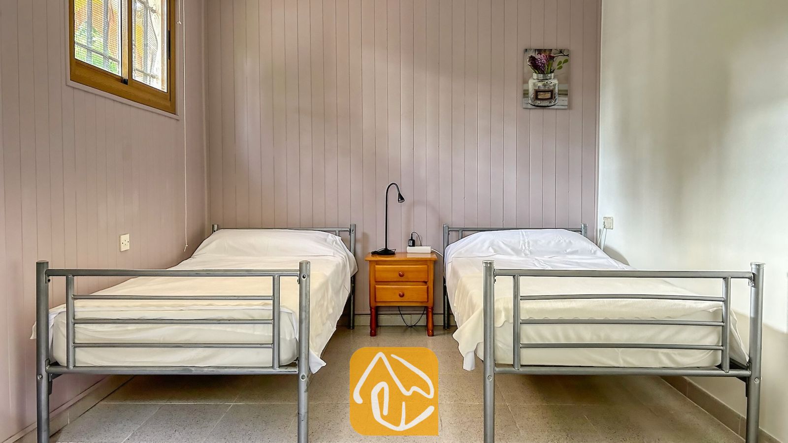 Vakantievilla's Costa Brava Spanje - Villa Iako - Slaapkamer met twee eenpersoonsbedden met grijze metalen frames en wit beddengoed, gescheiden door een houten nachtkastje met een zwarte lamp.