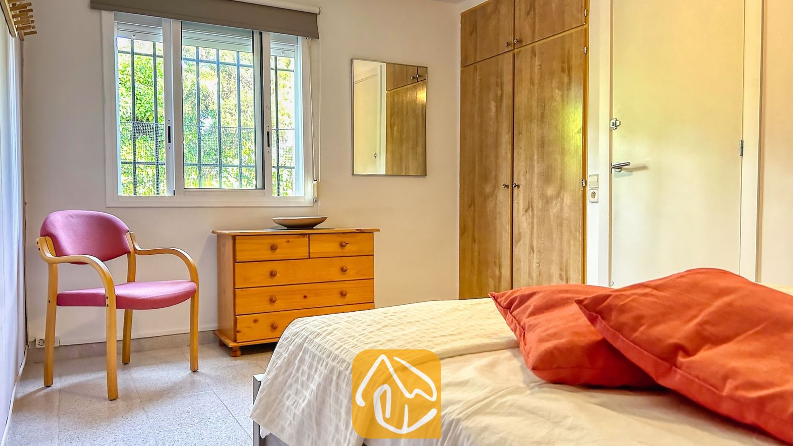 Vakantievilla's Costa Brava Spanje - Villa Iako - Slaapkamer met een bed voorzien van oranje kussens en een witte sprei, een houten ladekast en een roze fauteuil naast een raam met dievenklauwen.
