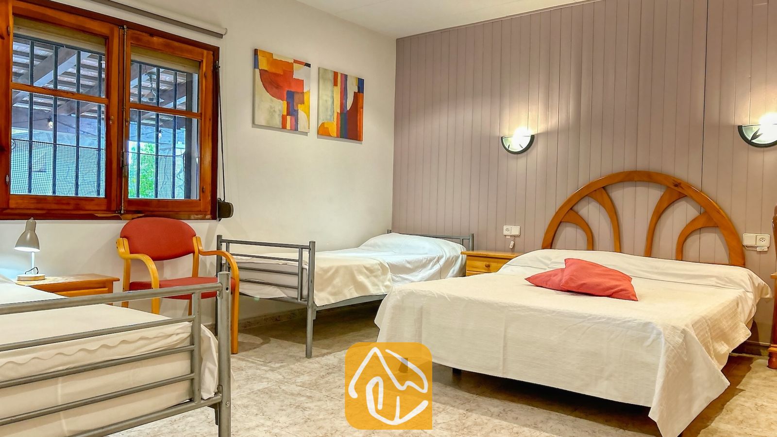 Vakantievilla's Costa Brava Spanje - Villa Iako - Slaapkamer in Villa Iako met een tweepersoonsbed voorzien van een houten hoofdbord, twee enkele bedden met een metalen frame, rode accentkussens en abstracte muurkunst.