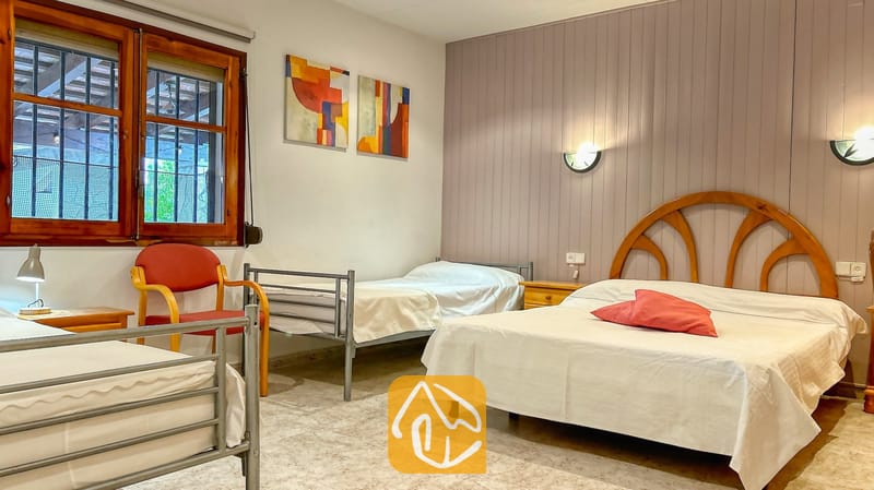 Villas de vacances Costa Brava Espagne - Villa Iako - Chambre de la Villa Iako dotée d'un lit double avec tête de lit en bois, de deux lits simples avec cadre en métal, de coussins décoratifs rouges et d'une œuvre d'art murale abstraite.