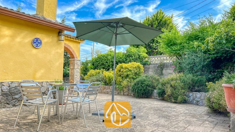 Vakantievilla's Costa Brava Spanje - Villa Iako - Eethoek in de buitenlucht bij Villa Iako, voorzien van metalen stoelen, een tafel met glazen blad en een grote groene parasol op een bestraat stenen terras.