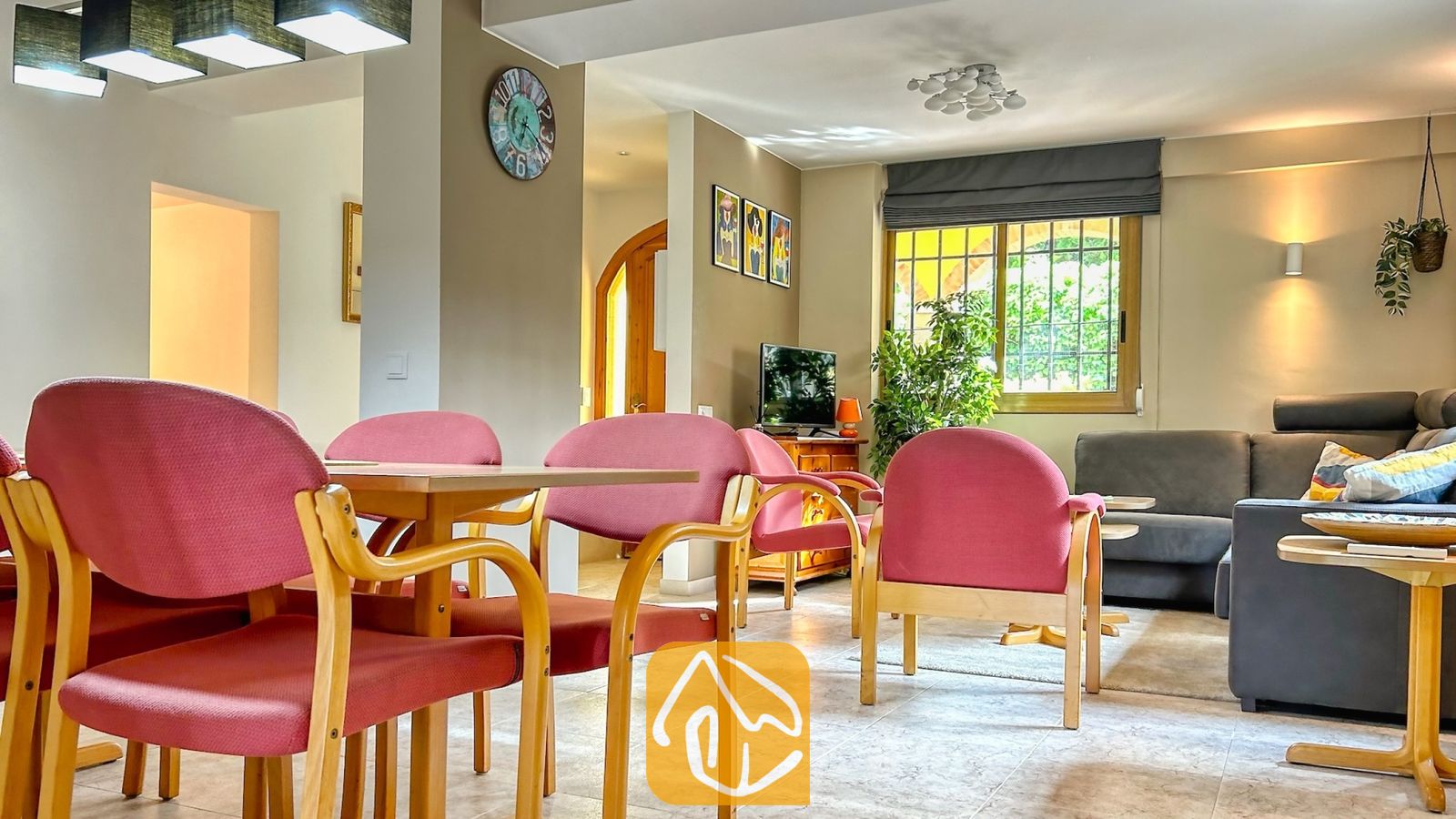 Vakantievilla's Costa Brava Spanje - Villa Iako - Open-plan woon- en eetgedeelte in Villa Iako, voorzien van houten fauteuils met roze kussens, een eettafel en een grijze hoekbank bij het raam.
