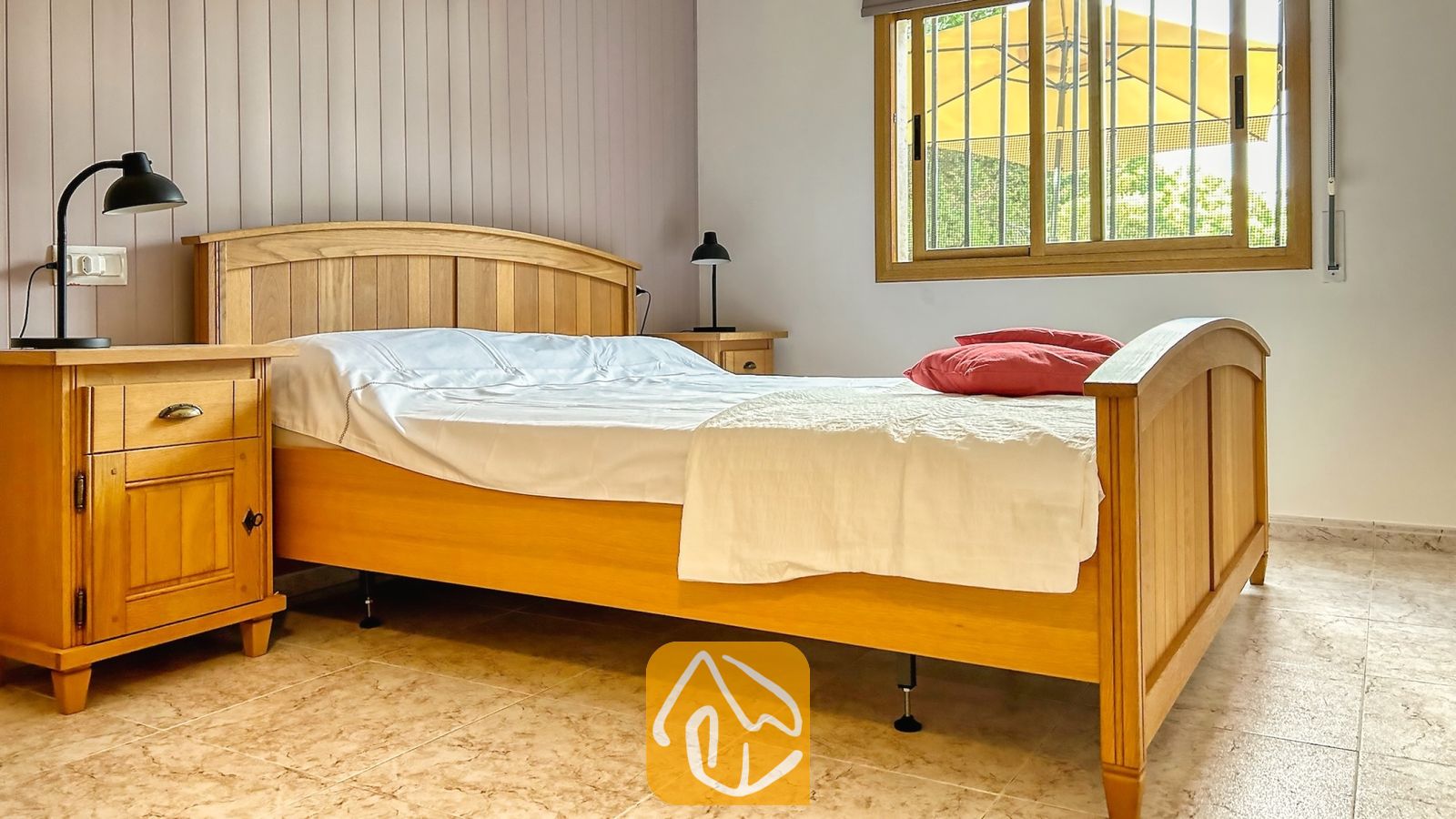 Vakantievilla's Costa Brava Spanje - Villa Iako - Slaapkamer met een houten bedframe, wit beddengoed, rode kussens en bijpassende houten nachtkastjes met zwarte lampen onder een raam.