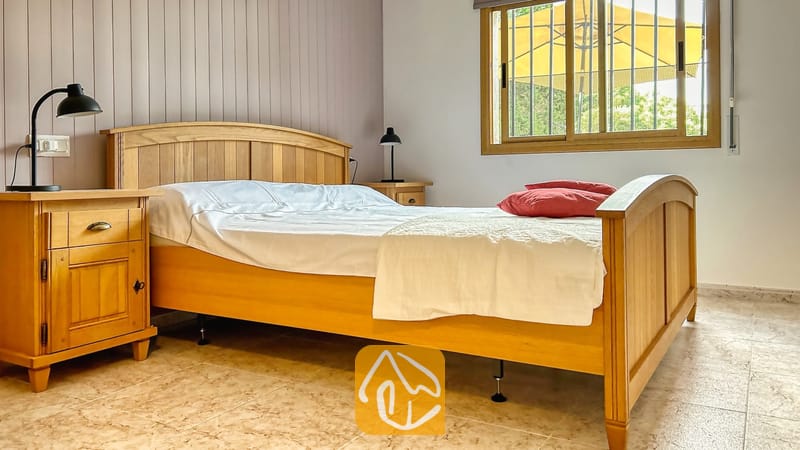 Villas de vacances Costa Brava Espagne - Villa Iako - Chambre avec un lit à cadre en bois, du linge de lit blanc, des coussins rouges et des tables de chevet en bois assorties surmontées de lampes noires, le tout sous une fenêtre.