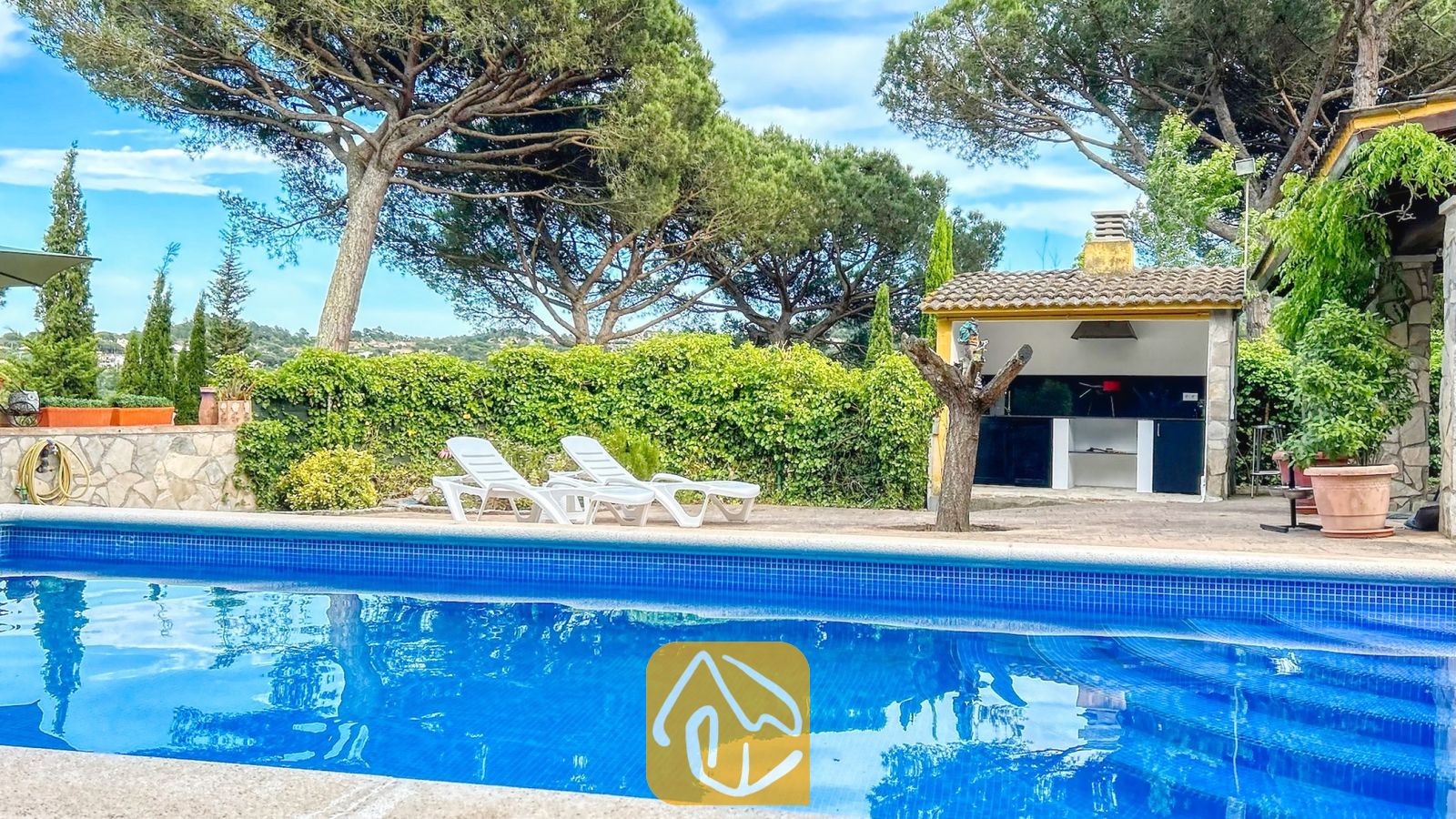 Vakantievilla's Costa Brava Spanje - Villa Iako - Privézwembad met blauw getegelde rand en witte ligstoelen op het terras van Villa Iako, voorzien van een buitenhuisje met barbecue en hoge dennenbomen op de achtergrond.