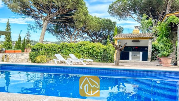 Vakantievilla's Costa Brava Spanje - Villa Iako - Privézwembad met blauw getegelde rand en witte ligstoelen op het terras van Villa Iako, voorzien van een buitenhuisje met barbecue en hoge dennenbomen op de achtergrond.