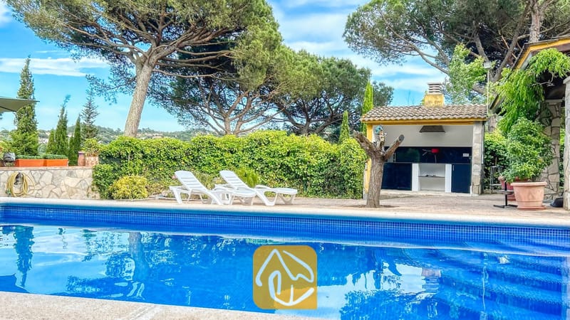 Vakantievilla's Costa Brava Spanje - Villa Iako - Privézwembad met blauw getegelde rand en witte ligstoelen op het terras van Villa Iako, voorzien van een buitenhuisje met barbecue en hoge dennenbomen op de achtergrond.