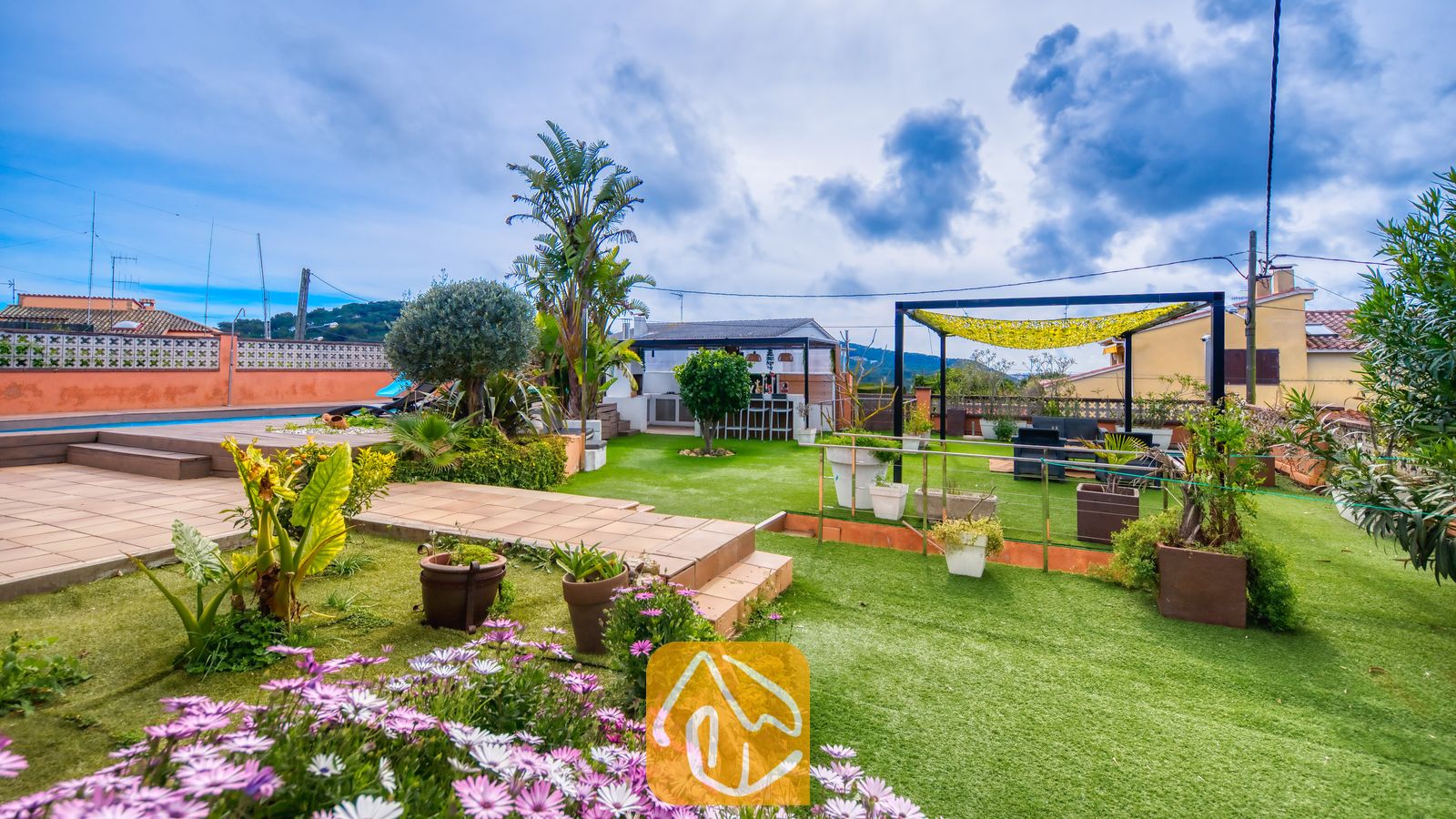 Villas de vacances Costa Brava Espagne - Villa Soleia - Terrasse extérieure de la Villa Soleia dotée de gazon synthétique, de fleurs mauves, de plantes en pots, d'un coin salon couvert et d'un bar extérieur sous un ciel bleu nuageux.
