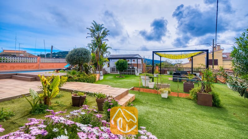 Villas de vacances Costa Brava Espagne - Villa Soleia - Terrasse extérieure de la Villa Soleia dotée de gazon synthétique, de fleurs mauves, de plantes en pots, d'un coin salon couvert et d'un bar extérieur sous un ciel bleu nuageux.