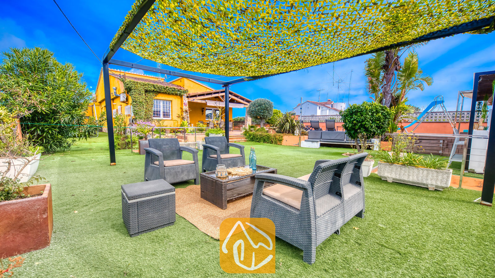 Villas de vacances Costa Brava Espagne - Villa Soleia - Espace salon extérieur à la Villa Soleia, comprenant des fauteuils en osier foncé, un canapé assorti et une table basse, installés sous une toile d'ombrage jaune sur une pelouse verdoyante.