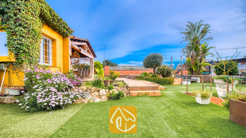 Villas de vacances Costa Brava Espagne - Villa Soleia - Jardin ensoleillé et mur extérieur jaune de la Villa Soleia, ornés d'une pelouse verte, de fleurs mauves et de palmiers sous un ciel bleu.