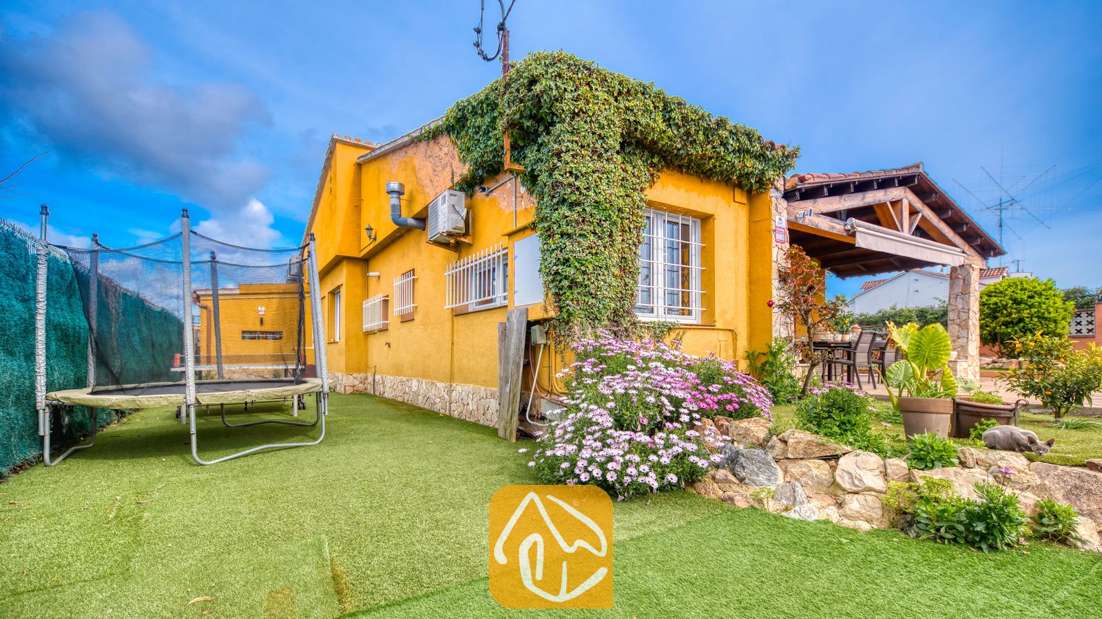 Villas de vacances Costa Brava Espagne - Villa Soleia - Vue extérieure de la Villa Soleia montrant une maison jaune couverte de lierre grimpant, un trampoline sur pelouse synthétique et une terrasse couverte.