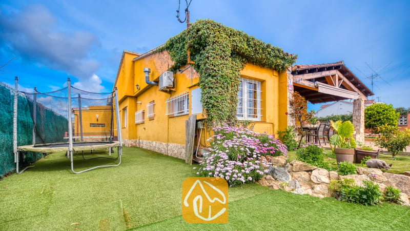 Villas de vacances Costa Brava Espagne - Villa Soleia - Vue extérieure de la Villa Soleia montrant une maison jaune couverte de lierre grimpant, un trampoline sur pelouse synthétique et une terrasse couverte.