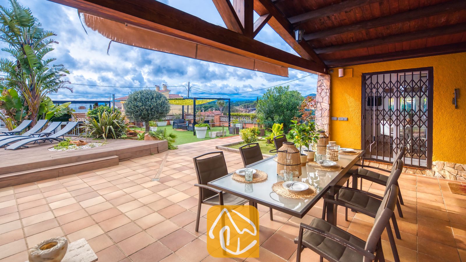 Villas de vacances Costa Brava Espagne - Villa Soleia - Terrasse extérieure couverte de la Villa Soleia comprenant une table avec plateau en verre, des chaises en métal marron et un sol carrelé, offrant une vue sur la terrasse du jardin et un ciel nuageux.