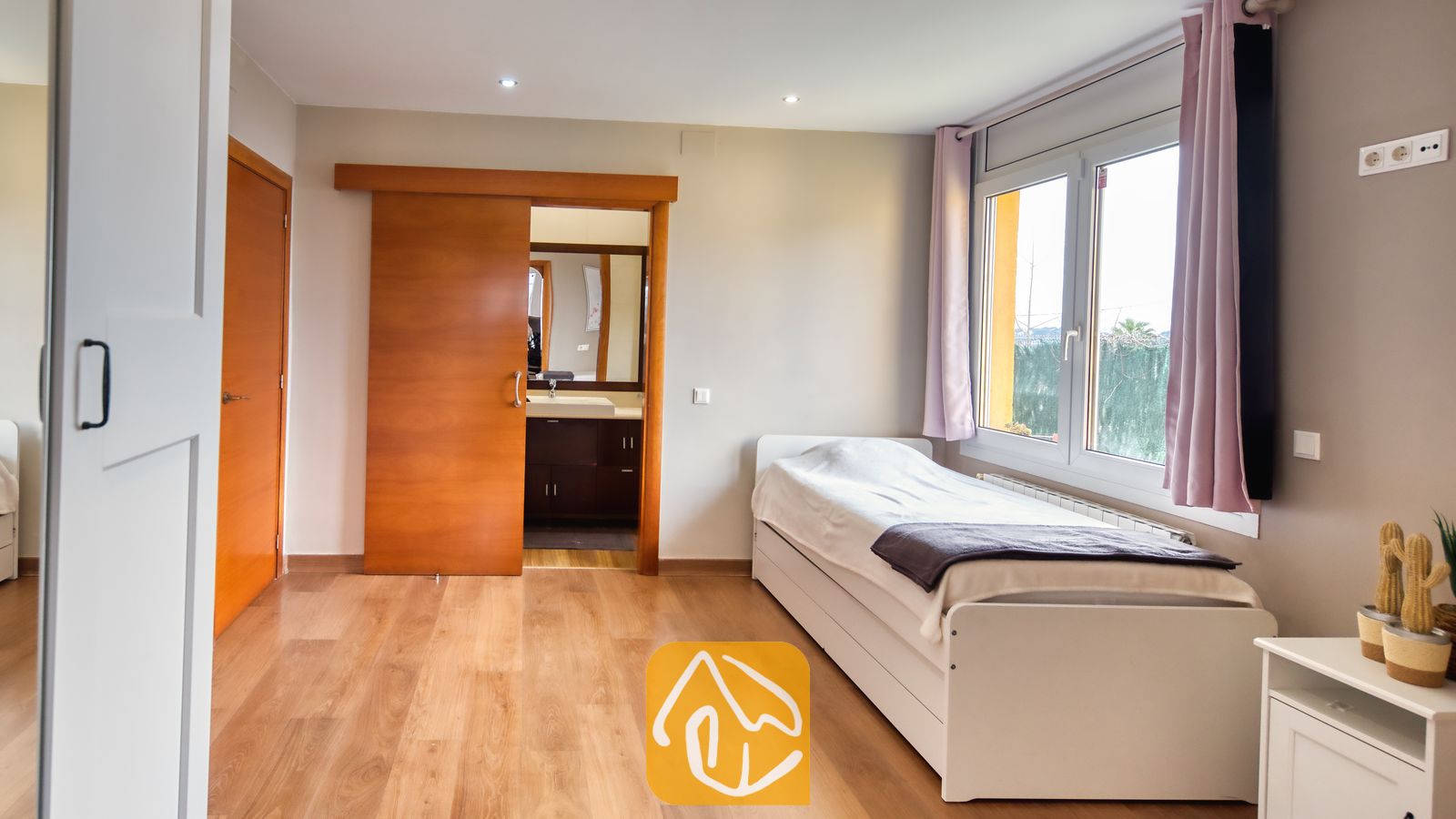 Villas de vacances Costa Brava Espagne - Villa Soleia - Chambre avec un lit simple blanc, un parquet en bois, une fenêtre aux rideaux roses et une porte coulissante en bois ouverte laissant entrevoir une salle de bains attenante.
