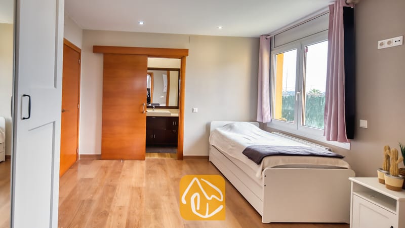 Villas de vacances Costa Brava Espagne - Villa Soleia - Chambre avec un lit simple blanc, un parquet en bois, une fenêtre aux rideaux roses et une porte coulissante en bois ouverte laissant entrevoir une salle de bains attenante.