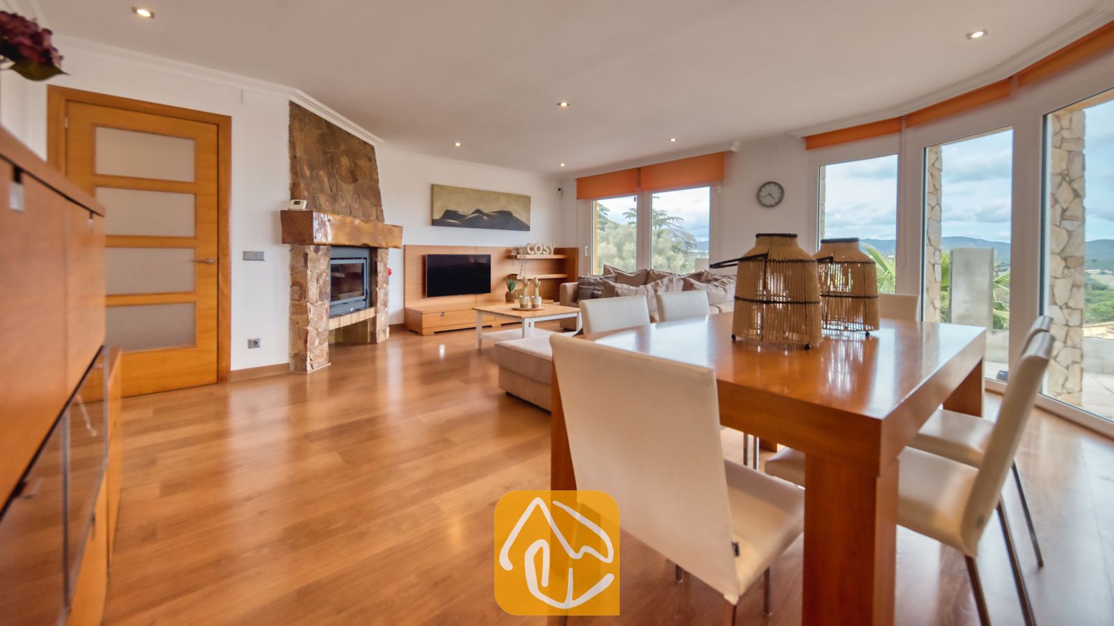 Villas de vacances Costa Brava Espagne - Villa Soleia - Spacieux salon et salle à manger à la Villa Soleia, comprenant une table à manger en bois, des chaises blanches, une cheminée en pierre et de grandes fenêtres.