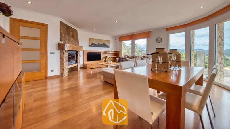 Villas de vacances Costa Brava Espagne - Villa Soleia - Spacieux salon et salle à manger à la Villa Soleia, comprenant une table à manger en bois, des chaises blanches, une cheminée en pierre et de grandes fenêtres.