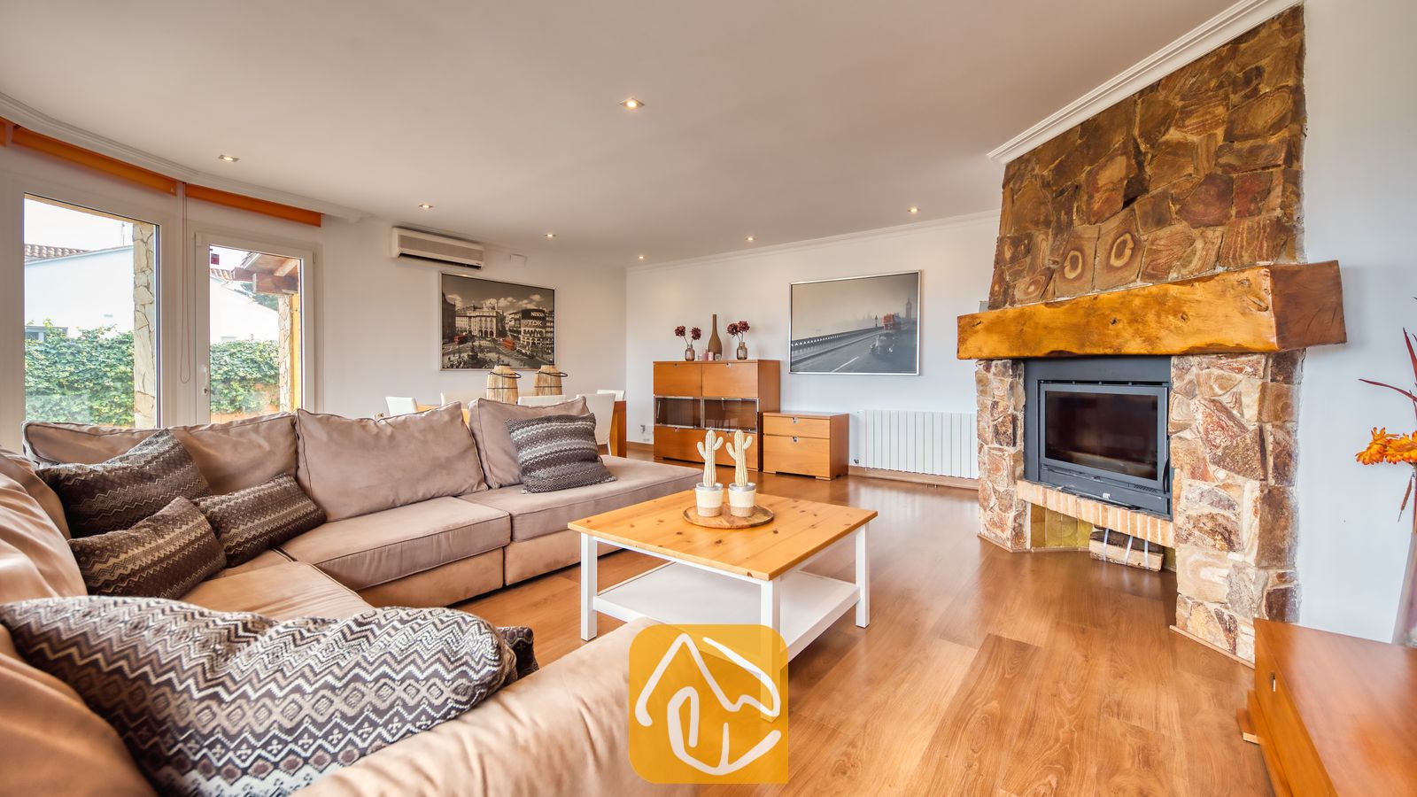 Villas de vacances Costa Brava Espagne - Villa Soleia - Spacieux salon de la Villa Soleia doté d'un grand canapé d'angle beige, d'une table basse en bois et d'une cheminée en pierre rustique surmontée d'un épais manteau de cheminée en bois.