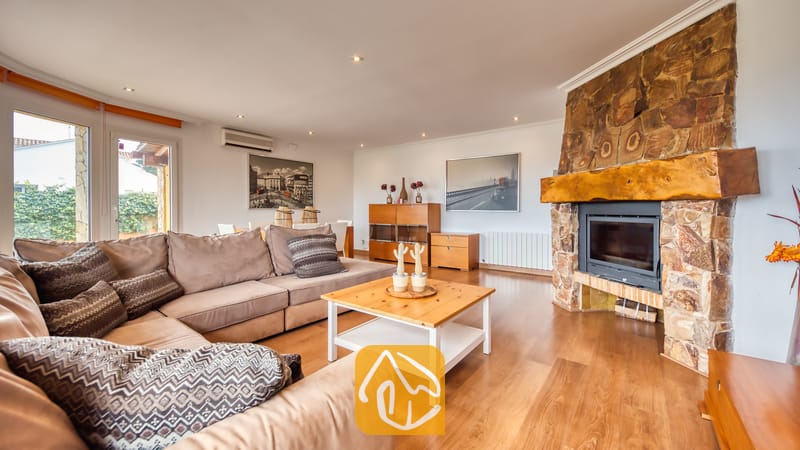 Villas de vacances Costa Brava Espagne - Villa Soleia - Spacieux salon de la Villa Soleia doté d'un grand canapé d'angle beige, d'une table basse en bois et d'une cheminée en pierre rustique surmontée d'un épais manteau de cheminée en bois.