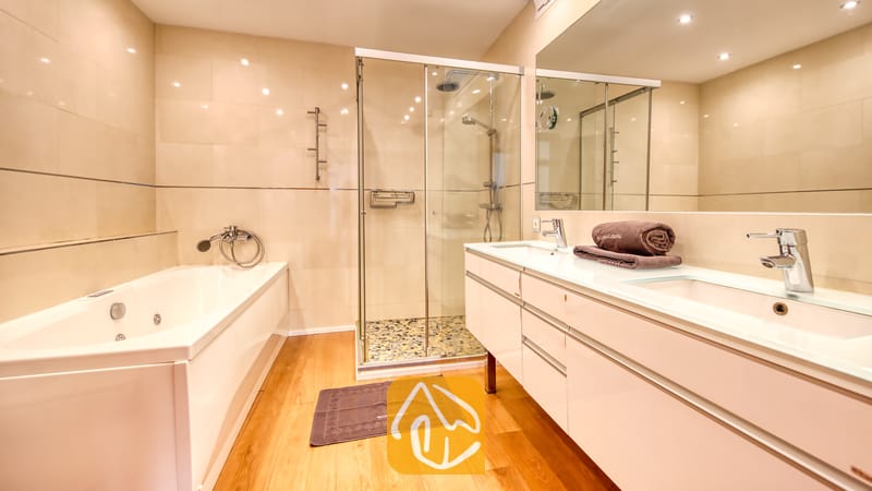 Villas de vacances Costa Brava Espagne - Villa Soleia - Salle de bains moderne comprenant une baignoire blanche, une douche à l'italienne vitrée, une double vasque avec des serviettes marron et des murs carrelés de couleur claire.